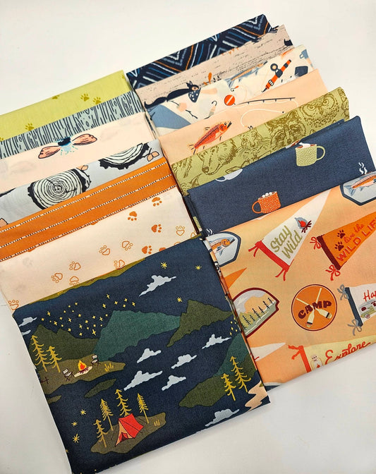 A curated 14-piece fabric bundle featuring Art Gallery Fabrics' Happy Camper and Little Forester collections. The fabrics are stacked neatly, showing off outdoor camping and woodland themes, including a dark navy main print with a campfire, tent, and starry mountain night sky, alongside coordinating prints of forest animals, fish, pennants, and tree bark textures in shades of navy, peach, olive green, and cream.
