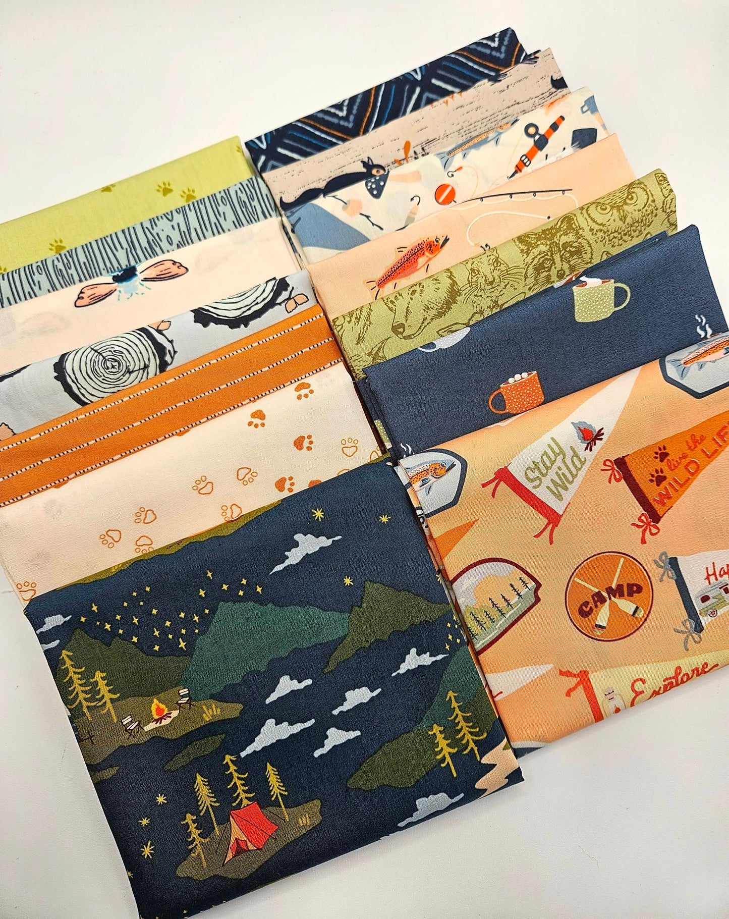 A curated 14-piece fabric bundle featuring Art Gallery Fabrics' Happy Camper and Little Forester collections. The fabrics are stacked neatly, showing off outdoor camping and woodland themes, including a dark navy main print with a campfire, tent, and starry mountain night sky, alongside coordinating prints of forest animals, fish, pennants, and tree bark textures in shades of navy, peach, olive green, and cream.
